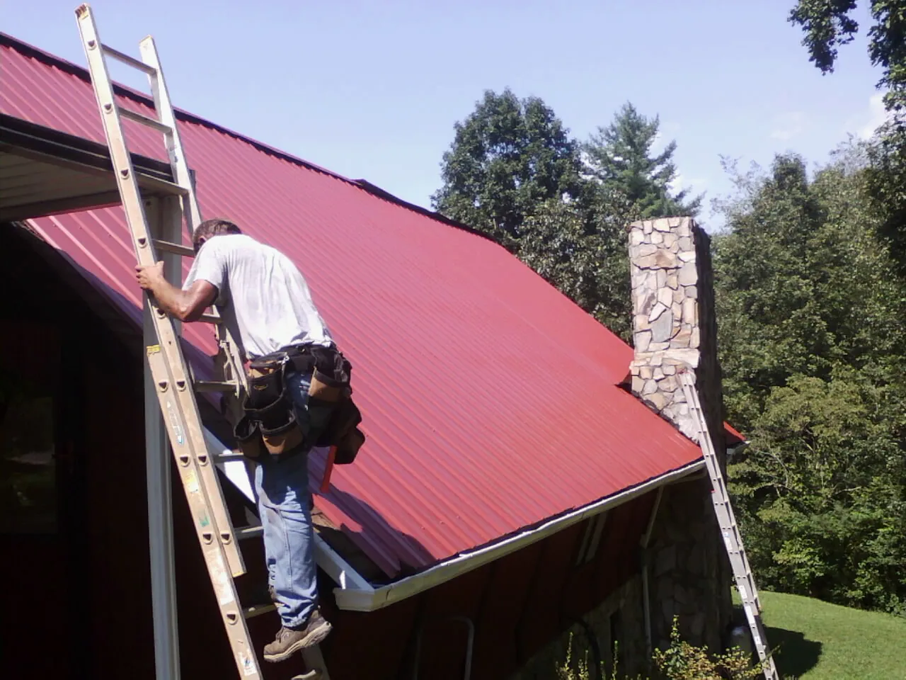 Roofer on a ladder beside a red metal roof and stone chimney — Soffit & Fascia Repair by Roofing Brothers in Briggsdale